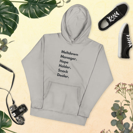 Gray hoodie with text 'Meltdown Manager, Hope Holder, Snack Dealer' on a beige background with plants and shoes.