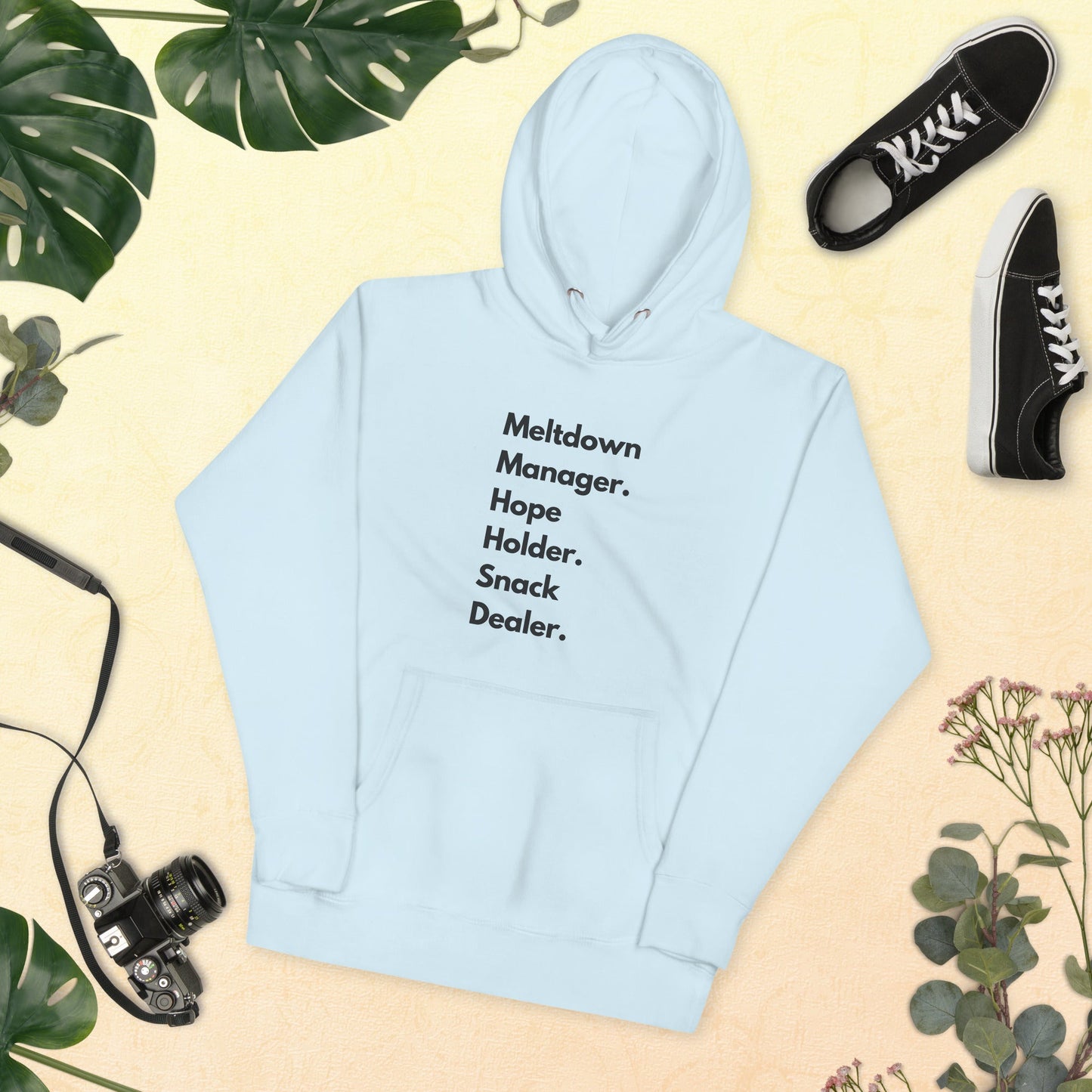 Light blue hoodie with text 'Meltdown Manager. Hope Holder. Snack Dealer.' on a beige background with plants and shoes.