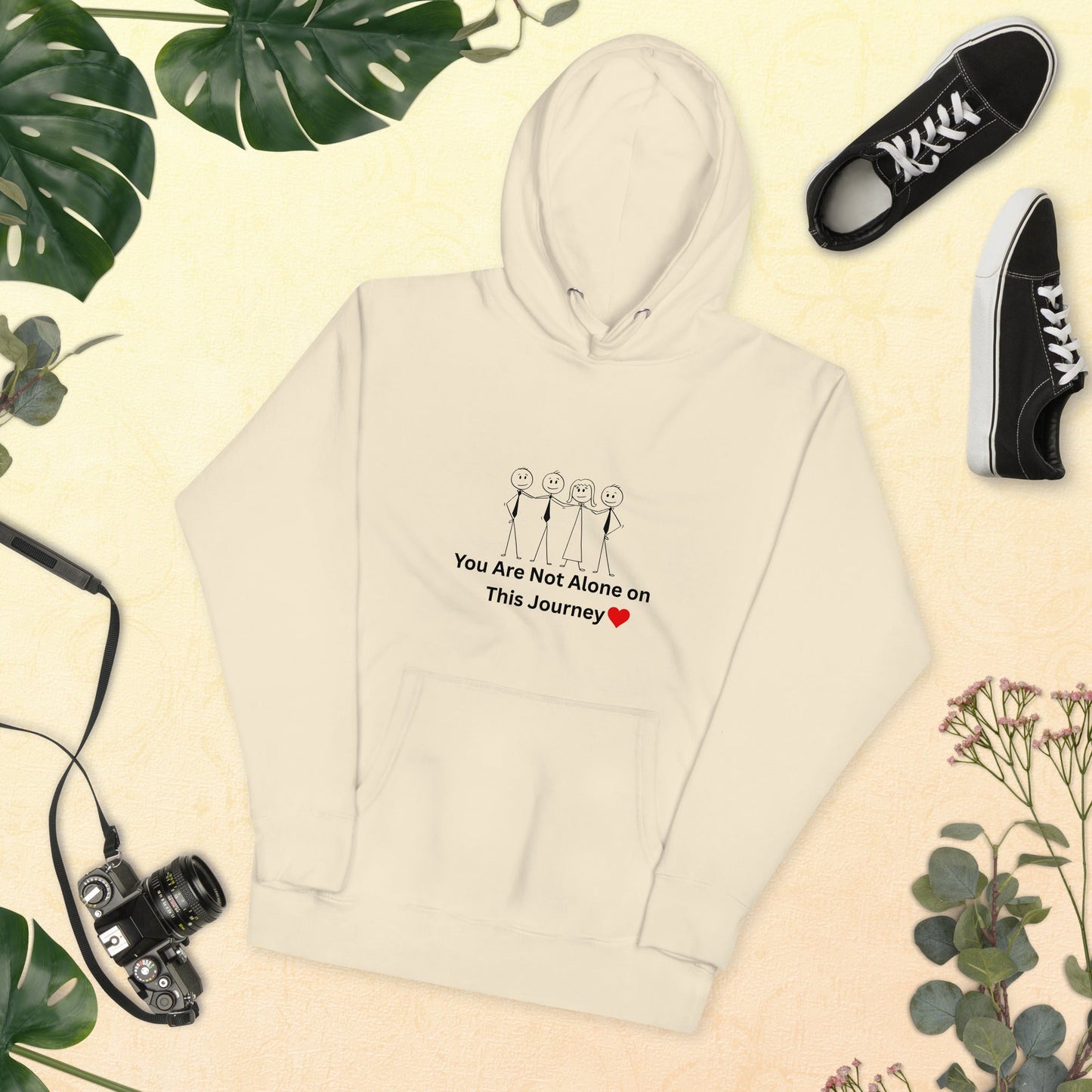 Beige hoodie with text and graphics, black sneakers, camera, and green leaves on a beige background